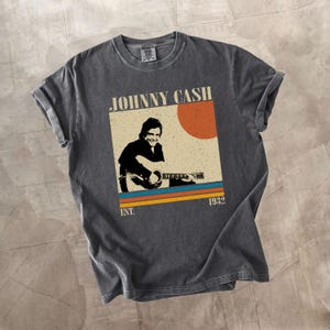 May include: Dark gray t-shirt featuring a vintage-style graphic of Johnny Cash playing guitar. The design includes the text "JOHNNY CASH" above an image of Cash, with a sun and stripes in orange, blue, and yellow. The text "EST. 1932" is below the image.