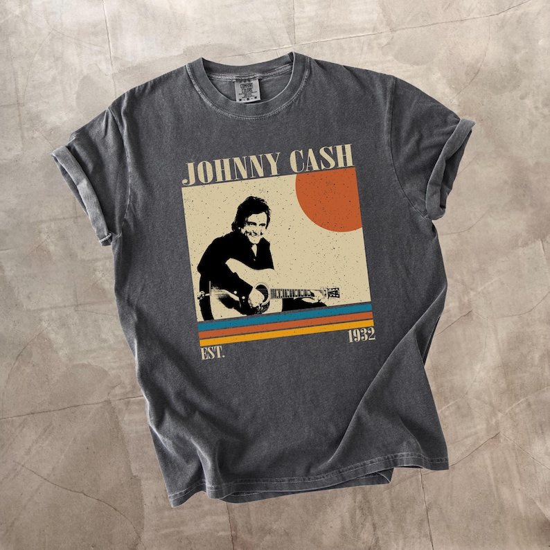 May include: Dark gray t-shirt featuring a vintage-style graphic of Johnny Cash playing guitar. The design includes the text "JOHNNY CASH" above an image of Cash, with a sun and stripes in orange, blue, and yellow. The text "EST. 1932" is below the image.