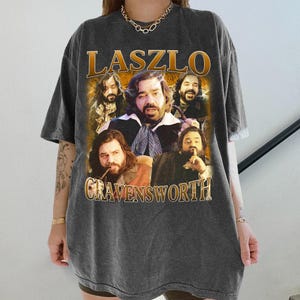 Könnte beinhalten: Dunkelgraues T-Shirt mit einem Grafikdesign im Vintage-Stil, das einen Mann mit langen Haaren und Bart zeigt. Die Grafik enthält den Namen "LASZLO" in goldfarbenen Buchstaben über dem Bild und "CRAVENSWORTH" darunter. Das T-Shirt hat ein leicht verwaschenes Aussehen.