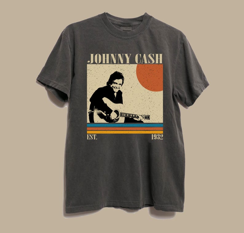 May include: Dark gray t-shirt featuring a graphic of Johnny Cash playing guitar. The design includes a retro-style image of Cash, a red sun, and the text "JOHNNY CASH" and "EST. 1932".