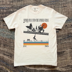 Puede incluir: Camiseta blanca con un gr&aacute;fico de la pel&iacute;cula "Spider-Man: Into the Spider-Verse". El dise&ntilde;o incluye siluetas de personajes, un paisaje urbano y el texto "EST. 2018". La camiseta tiene cuello redondo y mangas cortas.