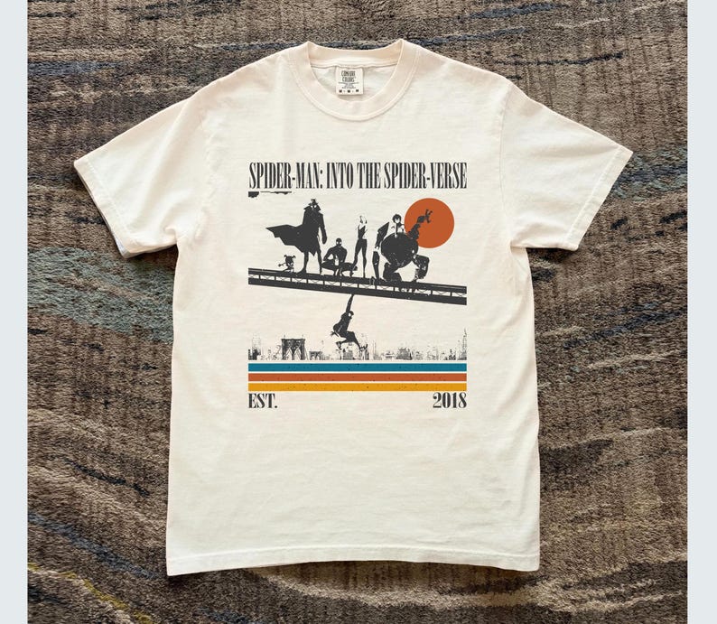 Puede incluir: Camiseta blanca con un gr&aacute;fico de la pel&iacute;cula "Spider-Man: Into the Spider-Verse". El dise&ntilde;o incluye siluetas de personajes, un paisaje urbano y el texto "EST. 2018". La camiseta tiene cuello redondo y mangas cortas.
