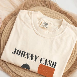 May include: Cream-colored t-shirt with the text "JOHNNY CASH" in black block letters. The shirt features a graphic of a person and a rust-colored rectangle. The shirt is folded on a woven, round placemat.