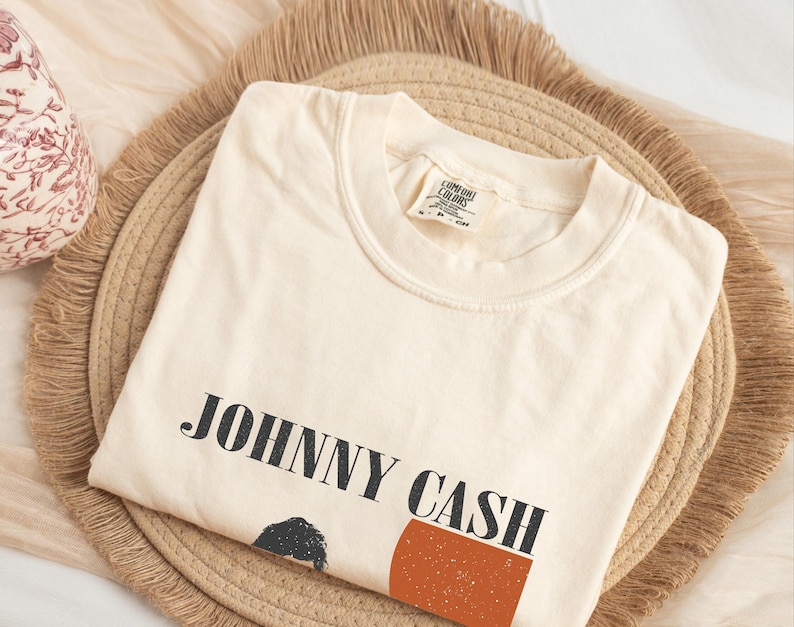 May include: Cream-colored t-shirt with the text "JOHNNY CASH" in black block letters. The shirt features a graphic of a person and a rust-colored rectangle. The shirt is folded on a woven, round placemat.