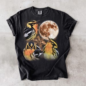 May include: A black short-sleeved t-shirt featuring a graphic print of three yellow and black birds with orange chests positioned around a large, textured, circular moon against a dark, cloudy background. The shirt has a crew neckline.