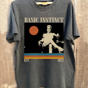 May include: Dark gray t-shirt featuring a graphic from the movie "Basic Instinct." The design includes the film's title, a retro image, and the text "EST. 1922" in a vintage style. The shirt is on a wooden hanger.
