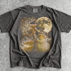 May include: A charcoal gray short-sleeve t-shirt featuring a graphic print of three textured toads positioned around a large, glowing yellow full moon. The shirt has a crew neckline and a faded, vintage-inspired wash throughout the fabric.