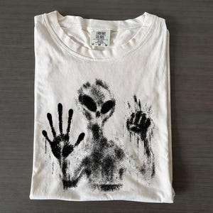 Op de afbeelding: Een gebroken wit t-shirt met korte mouwen en een zwarte, verweerde afbeelding van een alienfiguur met één hand tegen een oppervlak gedrukt. Het shirt heeft een Comfort Colors-merklabel in de nek met de maat M duidelijk afgedrukt.