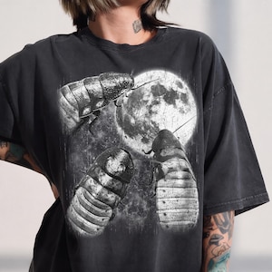 May include: A charcoal grey short-sleeve t-shirt featuring a graphic print of three large insects positioned around a textured, circular moon. The design is rendered in shades of black, white, and grey with a distressed, weathered appearance.
