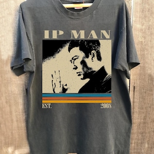 May include: Dark gray t-shirt featuring a graphic of a man with the text "IP MAN" above the image. Below the image are the words "EST. 2008" with a retro color stripe design. The shirt is displayed on a wooden hanger.