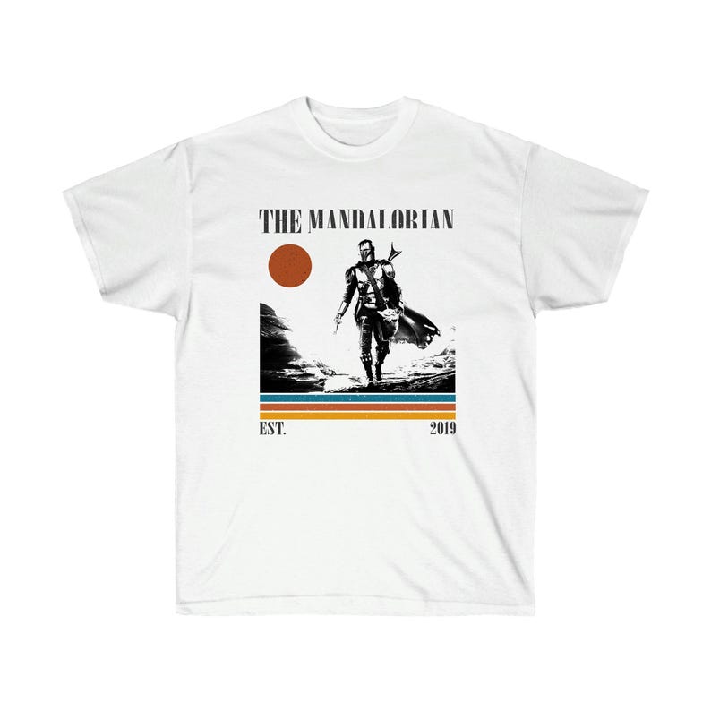 May include: White t-shirt featuring a black and white graphic of a figure from The Mandalorian series. The design includes a brown sun, blue and orange stripes, and the text "THE MANDALORIAN" above the image, with "EST. 2019" below.