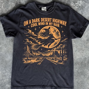 May include: A black short-sleeved t-shirt featuring an orange graphic of a witch flying on a broomstick in front of a full moon over a desert highway. The text reads: ON A DARK DESERT HIGHWAY COOL WIND IN MY HAIR. The design includes cacti and stars.