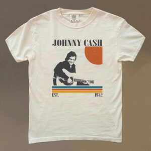 May include: Cream-colored t-shirt featuring a retro design with the text "JOHNNY CASH" above an illustration of Johnny Cash playing a guitar. The design includes a red-orange sun and horizontal stripes. The text "EST. 1932" is below the stripes.