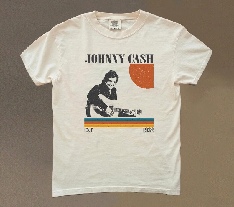 May include: Cream-colored t-shirt featuring a retro design with the text "JOHNNY CASH" above an illustration of Johnny Cash playing a guitar. The design includes a red-orange sun and horizontal stripes. The text "EST. 1932" is below the stripes.