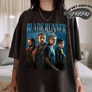 May include: Black t-shirt with a vintage-style graphic of the movie "Blade Runner 2049". The graphic features the main characters from the film, including Ryan Gosling, Harrison Ford, and Ana de Armas.