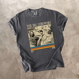 May include: Dark grey t-shirt with a graphic from the film "The Big Lebowski." The graphic shows three men, the film's title, and the year 1998. The shirt has rolled-up sleeves and a crew neck.