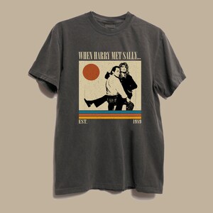 Puede incluir: Camiseta gris oscuro con un gr&aacute;fico de la pel&iacute;cula "Cuando Harry encontr&oacute; a Sally...". El dise&ntilde;o incluye una imagen retro de los dos personajes principales, un sol y el texto "EST. 1989".