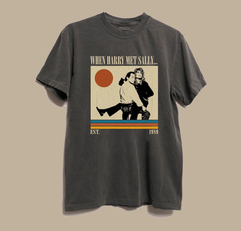 Puede incluir: Camiseta gris oscuro con un gr&aacute;fico de la pel&iacute;cula "Cuando Harry encontr&oacute; a Sally...". El dise&ntilde;o incluye una imagen retro de los dos personajes principales, un sol y el texto "EST. 1989".
