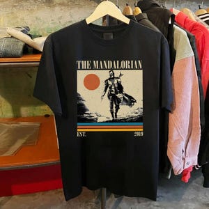 May include: Black t-shirt featuring a graphic of The Mandalorian. The design includes a figure walking on a planet, a sun, and the text "THE MANDALORIAN" and "EST. 2019". The shirt is hanging on a wooden hanger.