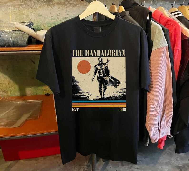 May include: Black t-shirt featuring a graphic of The Mandalorian. The design includes a figure walking on a planet, a sun, and the text "THE MANDALORIAN" and "EST. 2019". The shirt is hanging on a wooden hanger.