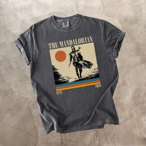 May include: Dark gray t-shirt featuring a graphic of The Mandalorian. The design includes an image of the character, a setting sun, and the text "THE MANDALORIAN" and "EST. 2019". The shirt has rolled-up sleeves.