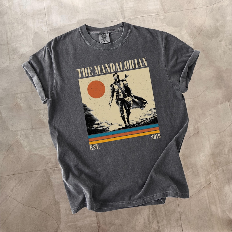 May include: Dark gray t-shirt featuring a graphic of The Mandalorian. The design includes an image of the character, a setting sun, and the text "THE MANDALORIAN" and "EST. 2019". The shirt has rolled-up sleeves.