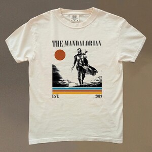 May include: Cream-colored t-shirt featuring a graphic of The Mandalorian. The design includes a figure walking across a landscape, a red sun, and the text "THE MANDALORIAN" above the figure. Below the figure are horizontal stripes and the text "EST. 2019".