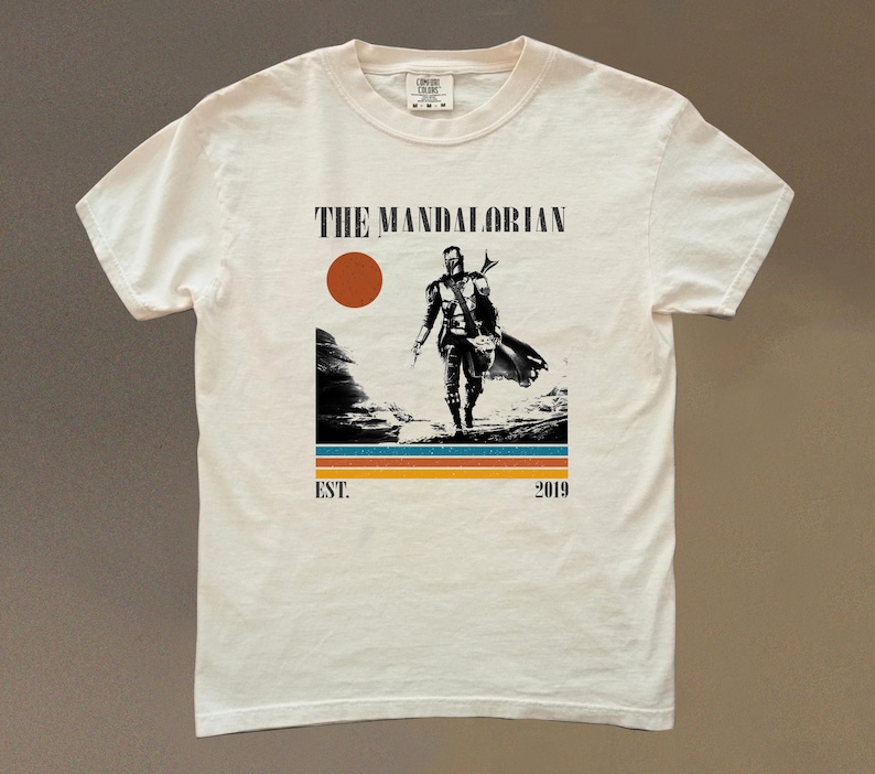 May include: Cream-colored t-shirt featuring a graphic of The Mandalorian. The design includes a figure walking across a landscape, a red sun, and the text "THE MANDALORIAN" above the figure. Below the figure are horizontal stripes and the text "EST. 2019".