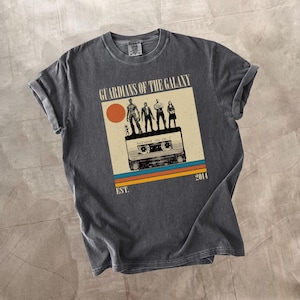 Może przedstawiać: Ciemnoszara koszulka z nadrukiem w stylu retro. Grafika zawiera napisy "GUARDIANS OF THE GALAXY" i "AWESOME MIX VOL. 1", z postaciami na kasecie, słońcem i wzorem w paski. Koszulka jest wstępnie skurczona.