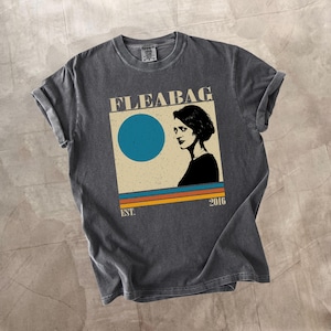 May include: Dark gray t-shirt featuring a graphic design with the word "FLEABAG", a blue circle, a woman's silhouette, and horizontal stripes in orange, yellow, and blue. The text "EST. 2016" is also included.