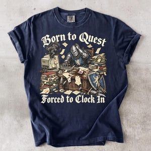May include: A navy blue t-shirt featuring a graphic of a knight in armor sitting at a desk with a computer, stacks of paper, and a shield. The text reads Born to Quest Forced to Clock In. The knight has a hand on their head near a smoking computer.