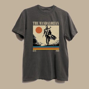 May include: Dark gray t-shirt featuring a graphic of The Mandalorian. The design includes a figure, a sunset, and the text "THE MANDALORIAN" above "EST. 2019". The shirt is a crew neck and has short sleeves.