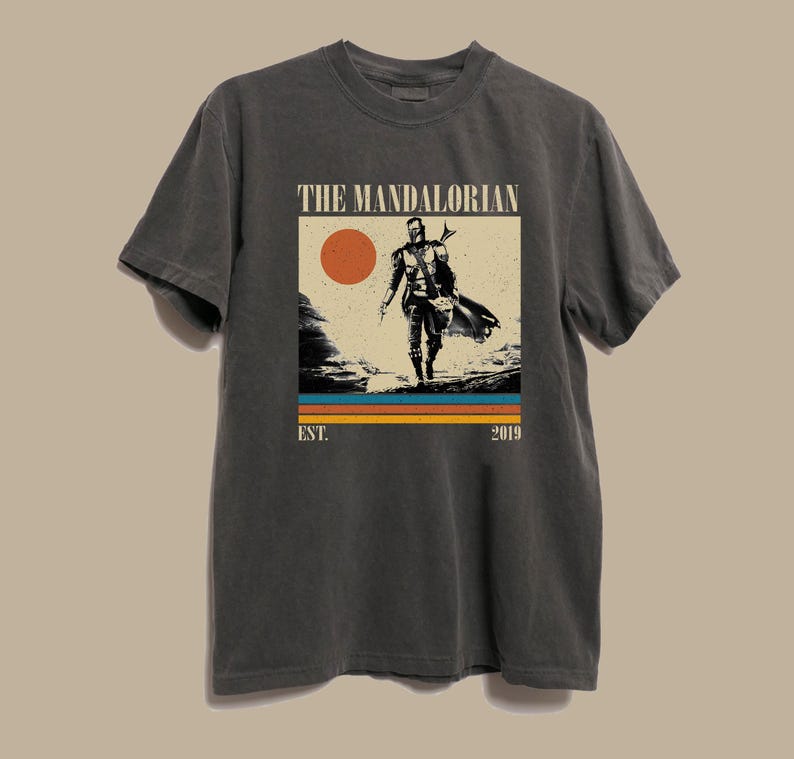 May include: Dark gray t-shirt featuring a graphic of The Mandalorian. The design includes a figure, a sunset, and the text "THE MANDALORIAN" above "EST. 2019". The shirt is a crew neck and has short sleeves.