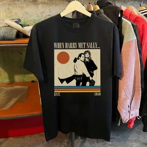 Puede incluir: Camiseta negra con un gr&aacute;fico de la pel&iacute;cula "Cuando Harry encontr&oacute; a Sally". El dise&ntilde;o incluye una ilustraci&oacute;n de dos personas, un sol y el texto "EST. 1989". La camiseta est&aacute; colgada en una percha de madera.