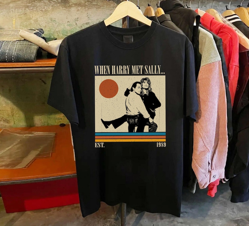Puede incluir: Camiseta negra con un gr&aacute;fico de la pel&iacute;cula "Cuando Harry encontr&oacute; a Sally". El dise&ntilde;o incluye una ilustraci&oacute;n de dos personas, un sol y el texto "EST. 1989". La camiseta est&aacute; colgada en una percha de madera.