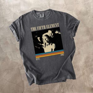 May include: Charcoal grey t-shirt with a graphic from the film "The Fifth Element." The design includes a movie still with the text "THE FIFTH ELEMENT" and "EST. 1997" in a retro style. The shirt has rolled-up sleeves.