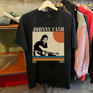 May include: Black t-shirt featuring a vintage-style graphic of Johnny Cash playing guitar. The design includes the text "JOHNNY CASH" above an image of Cash, with a retro sun and stripes. The shirt also has "EST. 1932" printed below the image.