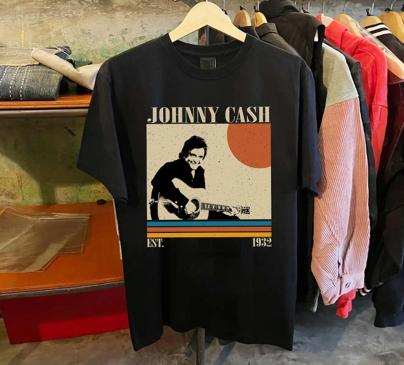 May include: Black t-shirt featuring a vintage-style graphic of Johnny Cash playing guitar. The design includes the text "JOHNNY CASH" above an image of Cash, with a retro sun and stripes. The shirt also has "EST. 1932" printed below the image.