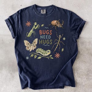 May include: A dark navy blue short-sleeve t-shirt featuring a graphic print of various insects including a praying mantis, a beetle, a moth, a caterpillar, and a stick insect, with the text "BUGS NEED HUGS" printed in the center in colorful letters.