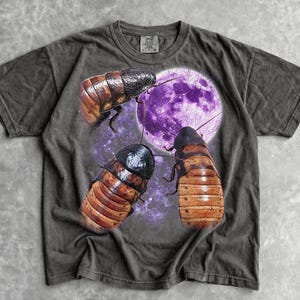 May include: A charcoal grey short-sleeve t-shirt featuring a graphic print of three brown cockroaches positioned around a large, glowing purple moon set against a starry space background. The shirt has a crew neckline and a visible brand label.