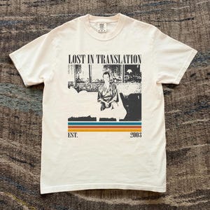 以下が含まれることがあります： 「LOST IN TRANSLATION」の文字と人物の白黒写真がプリントされたクリーム色のTシャツ。画像の下には水平ストライプと「EST. 2003」の文字があります。