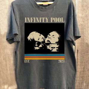 May include: Dark gray t-shirt with the words "INFINITY POOL" above a graphic of two faces in water. Below the graphic are horizontal stripes in blue, orange, and yellow, with "EST. 2023" below.