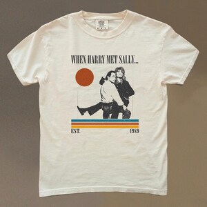 Puede incluir: Camiseta color crema con un gr&aacute;fico de la pel&iacute;cula "Cuando Harry encontr&oacute; a Sally...". El dise&ntilde;o incluye una imagen retro de dos personas, un c&iacute;rculo rojo y el texto "EST. 1989".