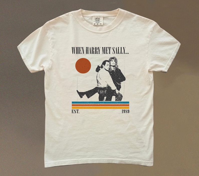 Puede incluir: Camiseta color crema con un gr&aacute;fico de la pel&iacute;cula "Cuando Harry encontr&oacute; a Sally...". El dise&ntilde;o incluye una imagen retro de dos personas, un c&iacute;rculo rojo y el texto "EST. 1989".