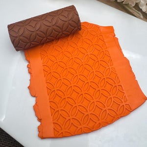 May include: A brown rolling pin with a geometric pattern and an orange textured sheet with the same pattern. The rolling pin is used to create an embossed design on the sheet.