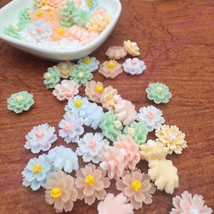 May include: A collection of pastel-colored, miniature flower embellishments. The flowers are in shades of pink, yellow, blue, green, and white, with yellow centers. Some are scattered on a brown surface, while others are in a white bowl.