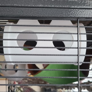 Peut inclure: Un accessoire blanc en plastique pour habitat d'animaux de compagnie avec deux ouvertures circulaires. En dessous, un hamac vert et marron pour animaux de compagnie est visible. L'accessoire est monté à l'intérieur d'une cage métallique.
