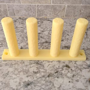 Peut inclure: Un support jaune pâle avec quatre poteaux cylindriques fixés à une base rectangulaire. Le support est conçu pour contenir des objets. Il est posé sur une surface mouchetée gris clair.
