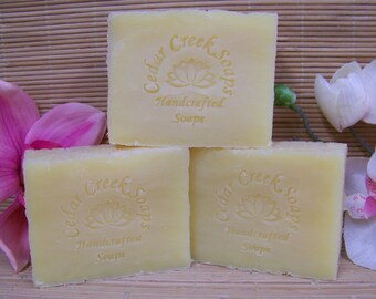 May include: Three bars of Cedar Creek Soaps handcrafted soap. The soap is a pale yellow color and has a floral design on the front. The soap is sitting on a bamboo mat with pink and white orchids.