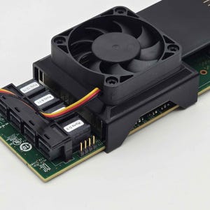 May include: Close-up of a black computer fan attached to a green circuit board. The fan has multiple blades and is mounted on a black housing. Several ports are labeled with white text. A multi-colored cable connects the fan to the board.